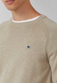 Mężczyzna w beżowym swetrze z dzianiny z żebrowanym kołnierzem i małym kwadratowym logo na piersi, mający na sobie białą koszulę pod spodem, eksponujący szyję i linię żuchwy.