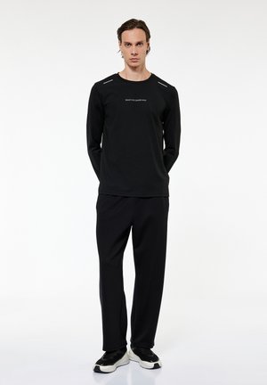 Mężczyzna stoi z rękami za plecami, ubrany w czarną koszulkę z długim rękawem z napisem „READY FOR CHANGE NOW”, czarne spodnie i czarno-białe sneakersy.