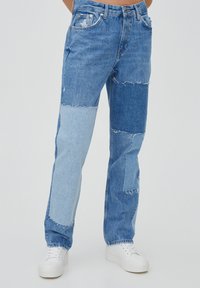 Jean en denim avec un design patchwork en nuances de bleu clair et foncé, bords effilochés, coupe droite, associé à des baskets blanches.