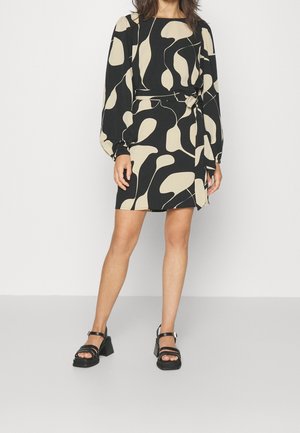 Vrouw draagt een mini-jurk met lange mouwen, een zwart-beige abstract patroon, een strikceintuur in de taille en zwarte sandalen met blokhak.