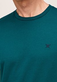 Muž v tyrkysovém tričku s kulatým výstřihem a malou vyšitou emblematickou značkou zkřížených golfových holí a golfového míčku na levé hrudi.