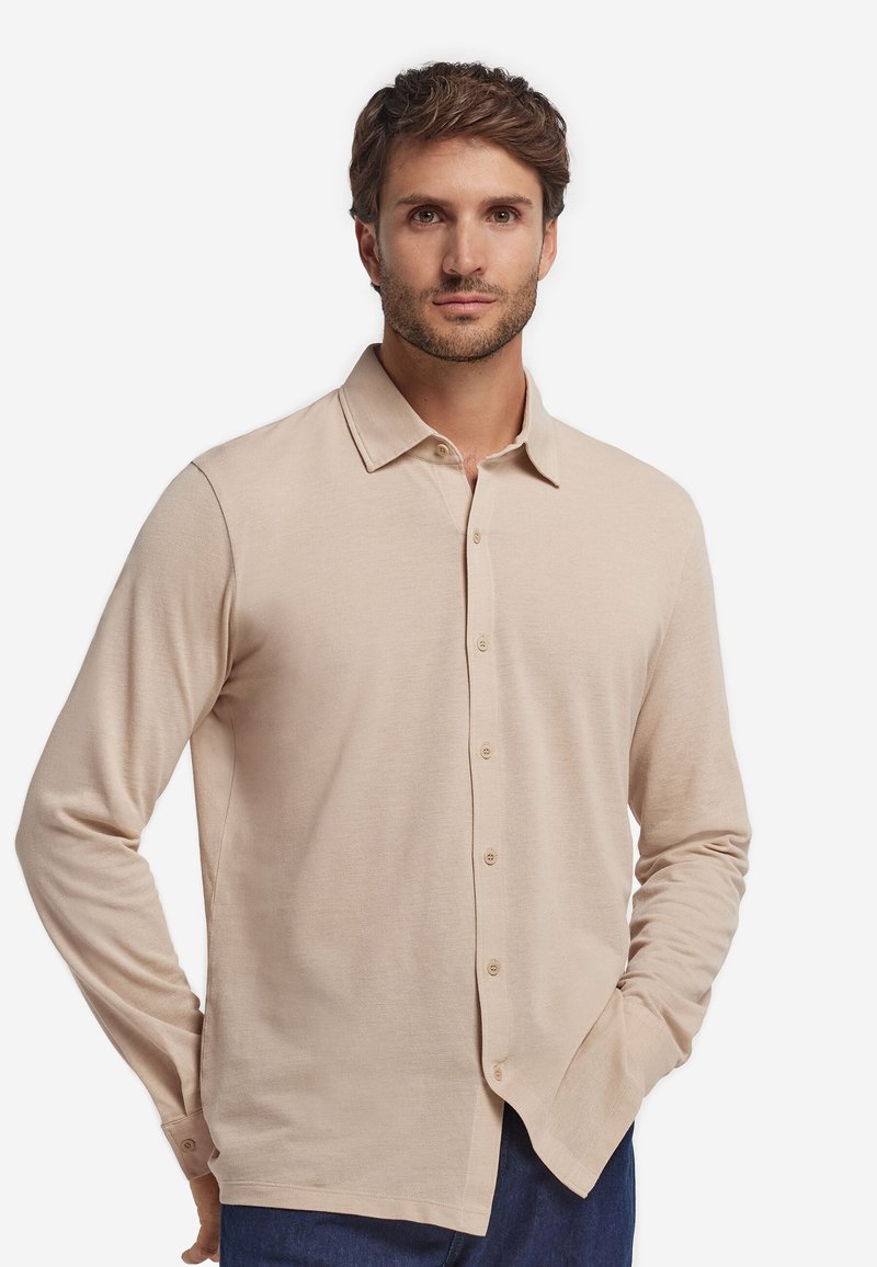 Mann mit kurzen braunen Haaren, trägt ein beiges langärmliges Hemd und blaue Hose, steht mit einer Hand in der Tasche vor weißem Hintergrund.