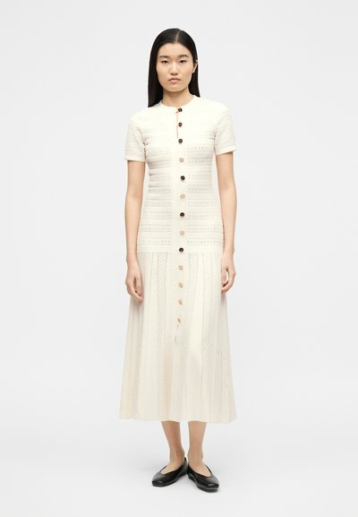 Γυναίκα που στέκεται όρθια φορώντας εκρού, midi φόρεμα με κοντά μανίκια και κουμπιά, και μαύρα μπαλαρίνες, σε λευκό φόντο.