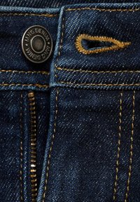 Tela de mezclilla azul oscuro con una cremallera de latón y un botón negro que presenta el logo "Ming Denim". Detalles de costura naranja a lo largo de las costuras.