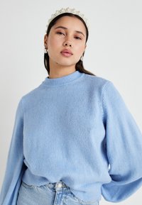Lys blå strikkegenser med høy krage og vide ermer, kombinert med lys denim. Stoffet er mykt og litt løst.