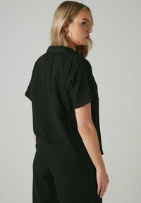 Maglietta nera a maniche corte con finitura testurizzata, design con colletto e vestibilità comoda; indossata da una persona con il corpo leggermente girato.