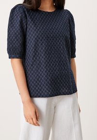 Blusa testurizzata blu navy con maniche a palloncino, caratterizzata da un motivo floreale e un colletto rotondo, abbinata a pantaloni larghi di colore chiaro.
