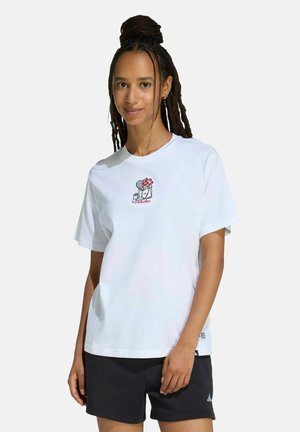 Mujer con el cabello largo y trenzado lleva una camiseta blanca con un pequeño estampado floral y de objetos y pantalones cortos negros sobre un fondo liso.
