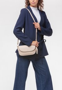 Borsa a tracolla beige con finitura in pelle liscia, pattina curva e tracolla regolabile, esposta su uno sfondo neutro.
