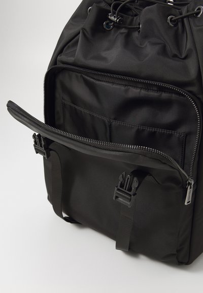 Musta reppu, joka on valmistettu kestävästä nylonista. Siinä on vetoketjullinen etutasku, solkiremmipäät sekä kiristysnyöri ylhäällä.