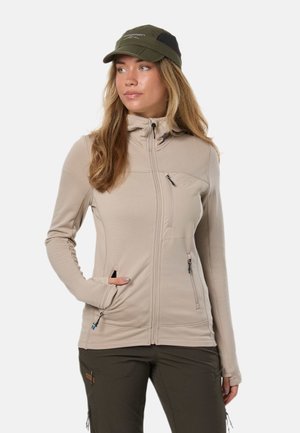 Mujer que lleva una chaqueta beige con cremallera y pantalones verde oliva con una gorra a juego, de pie con una mano en el bolsillo, mirando hacia un lado.