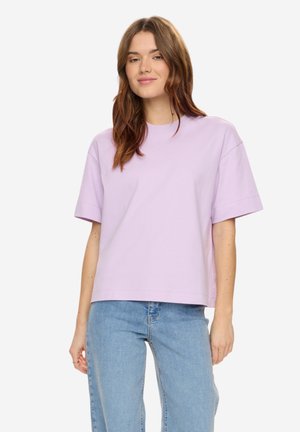 Frau mit hellbraunem, welligem Haar, die ein locker sitzendes, kurzärmeliges lavendelfarbenes Hemd und hellblaue Jeans trägt, stehend vor einem weißen Hintergrund.