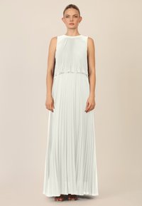Maxi-jurk van lichtgrijze stof met verticale plooien, mouwloos ontwerp, gelaagde bovenkant en vloeiende onderkant. Sandalen met zichtbare bruine banden.