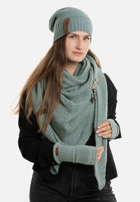 Mintgroene gebreide beanie met een bruin leren patch, bijpassende oversized sjaal en vingerloze handschoenen, gecombineerd met een zwarte trui.