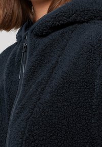 Svart fleecehuva med en mjuk, texturerad yta, framficka med dragkedja och justerbar dragsko på huvan.