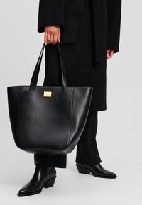 Borsa tote in pelle nera con una forma strutturata, caratterizzata da due manici lunghi e un dettaglio del logo in oro embossato sul davanti.