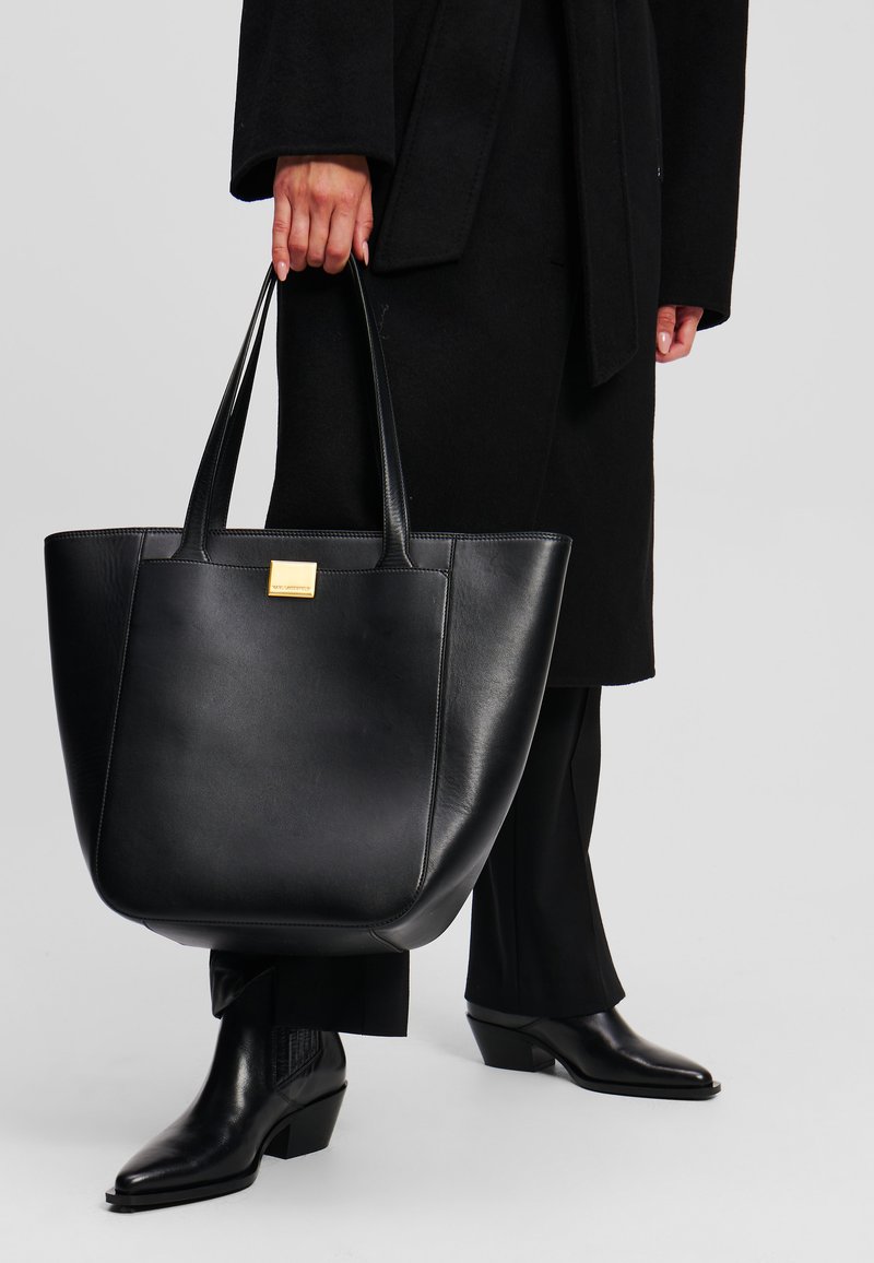 Borsa tote in pelle nera con una forma strutturata, caratterizzata da due manici lunghi e un dettaglio del logo in oro embossato sul davanti.