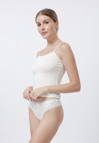 Vrouw in witte camisole en lingerie met kanttegenstaande tegen een effen witte achtergrond, kijkend naar de camera.