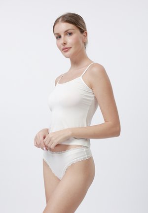 Woman in white camisole and lace-trimmed white underwear standing against a plain white background, looking at the camera.