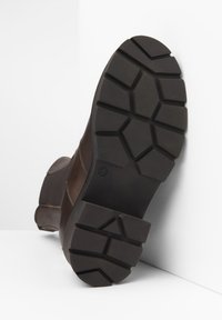 Botte en cuir marron avec une semelle en caoutchouc texturée arborant un motif de crampons géométrique. L'orteil est arrondi et il y a des détails de couture contrastants.