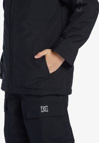 Svart vattentät jacka med dragkedja, sidofickor, justerbara ärmslut och DC-logotyp i grått på ärmen och fickan på byxorna.