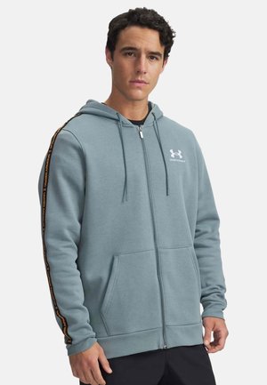 Homme portant un sweat à capuche zippé bleu Under Armour avec des bandes noires et orange de la marque sur les manches, debout devant un fond blanc.