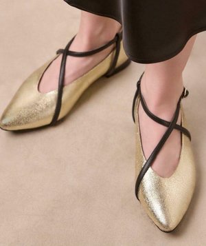 Bailarinas metálicas doradas con punta afilada y tiras cruzadas negras presentan una superficie texturizada y se encuentran sobre un fondo claro.