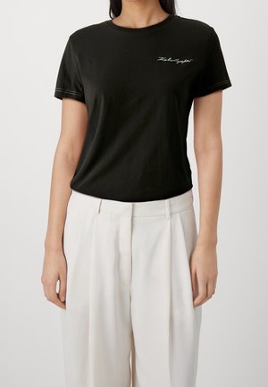 Kobieta w dopasowanym czarnym t-shircie z krótkim rękawem z małym białym napisem i luźnych białych spodniach z wysokim stanem, stojąca na jednolitym tle.