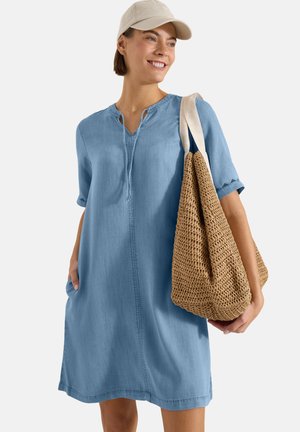 Kvinde iført lys blå løs kjole og beige kasket, der holder en stor flettet tan taske over den ene skulder, smilende og kiggende mod højre.