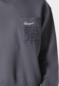 Sudadera gris hecha de una tela suave, con un bolsillo frontal que presenta un logotipo y una escritura en relieve, estilizada, en azul oscuro.