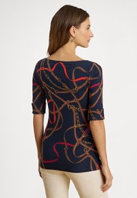 Ženska z rjavimi srednje dolgimi lasmi, oblečena v temno modro majico z vzorci rdečih in rjavih verig ter pasov, v kombinaciji z bež hlačami, stoji bočno.