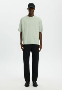 T-shirt ample vert clair à manches courtes, associée à un jean noir à jambes droites et des chaussures noires ; le mannequin porte une casquette noire.