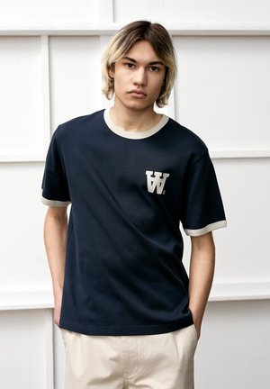 Marineblaue T-Shirt mit kontrastierendem cremefarbenem Kragen und Ärmeln; ausgestattet mit einem weißen Grafik-Logo auf der Brust; Baumwollmaterial.