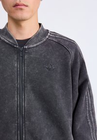 Casaco preto com fecho éclair, feito de tecido macio com uma textura desgastada. Apresenta um colarinho alto e um pequeno logótipo bordado no peito.