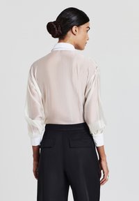 Una donna con i capelli scuri raccolti in uno chignon indossa una camicetta bianca trasparente a maniche lunghe con polsini bianchi e pantaloni neri a vita alta, rivolta di spalle alla macchina fotografica.