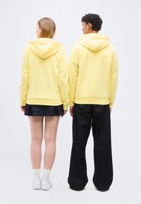 Deux personnes vues de dos portant des sweat-shirts à capuche jaune pâle, l'une avec une jupe noire et des baskets blanches, l'autre avec un pantalon large noir.