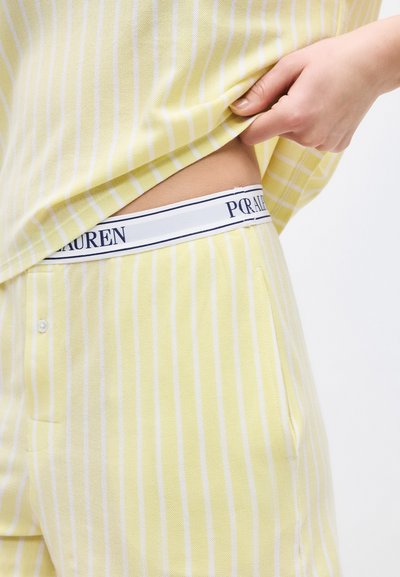 Το χέρι σηκώνει κίτρινο και λευκό ριγέ πουκάμισο, αποκαλύπτοντας μερικώς τη ζώνη κίτρινου ριγέ παντελονιού και λευκή ελαστική ταινία με μερικό κείμενο.