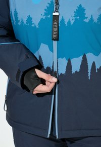 Ruka rozepínající modrou bundu s motivem hor a stromů, na ruce má černou rukavici bez prstů.