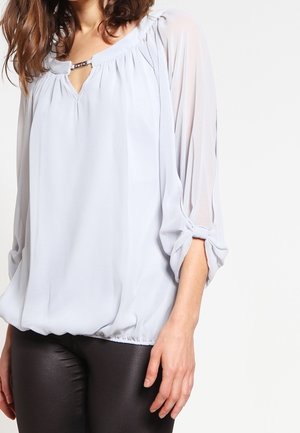 Frau trägt eine lockere, hellgraue Bluse mit transparenten 3/4-Ärmeln und einem Metallstabendetail am Ausschnitt, kombiniert mit schwarzer Hose.