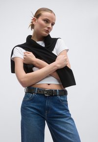 Black wrap, short-sleeved white crop top, high-waisted blue jeans, studded black belt. Smooth fabric, relaxed fit, minimalistic design.