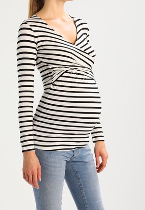 Donna incinta che indossa un top a maniche lunghe a righe bianche e nere e jeans azzurri, in piedi davanti a uno sfondo semplice.