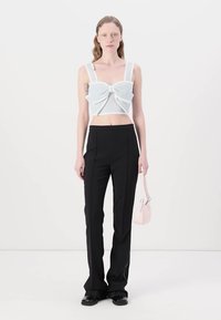 Crop top en tricot blanc avec un grand nœud, associé à un pantalon noir évasé taille haute. Le mannequin tient un petit sac à main rose clair.