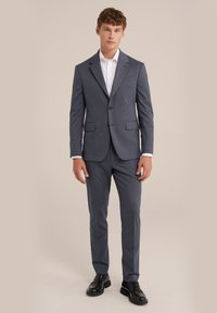 Grijze two-piece suit met een gestructureerd patroon, notch-lapels en een enkele knoopsluiting. Gecombineerd met zwarte dresschoenen en een wit shirt.