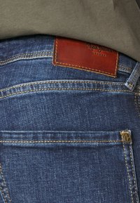 Mörkblå denimjeans med synlig sömnad, en brun läderlapp märkt "Pepe Jeans London", och en framficka med en logoknapp.