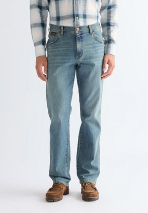 Mężczyzna w jasnoniebieskiej koszuli w kratę, prostych niebieskich dżinsach i brązowych skórzanych butach sznurowanych, stojący na jednolitym tle.