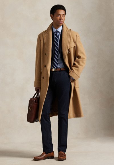 Muž v béžovém kabátě, tmavě modrém obleku, pruhované kravatě, hnědém pásku a botách, držící hnědou koženou aktovku, stojící před neutrálním pozadím.