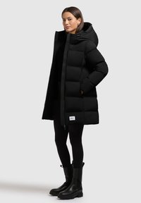 Abrigo acolchado negro con capucha, con un diseño de acolchado, cierre de cremallera y una etiqueta de marca en el dobladillo. Llevado con leggins negros y botas.