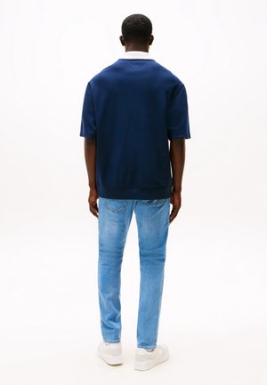 Mann steht mit dem Rücken zur Kamera, trägt ein marineblaues Kurzarmhemd, hellblaue Jeans und weiße Turnschuhe vor weißem Hintergrund.