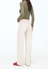 Olijfgroene long-sleeve top gecombineerd met hoge crèmekleurige wide-leg broeken en rode sneakers, die een getailleerd silhouet en een soepele textuur presenteren.