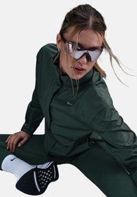 Žalia lengva striukė su aukštu apykakle ir logotipu, derinama su atitinkančiomis legginsais, juodais ir baltais sportbačiais bei veidrodiniais akiniais nuo saulės.